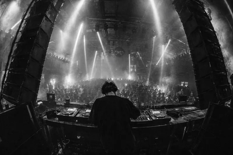 Black and white wide shot from behind a DJ at a large desk on stage, between two leaning speaker stacks, light beams in haze.