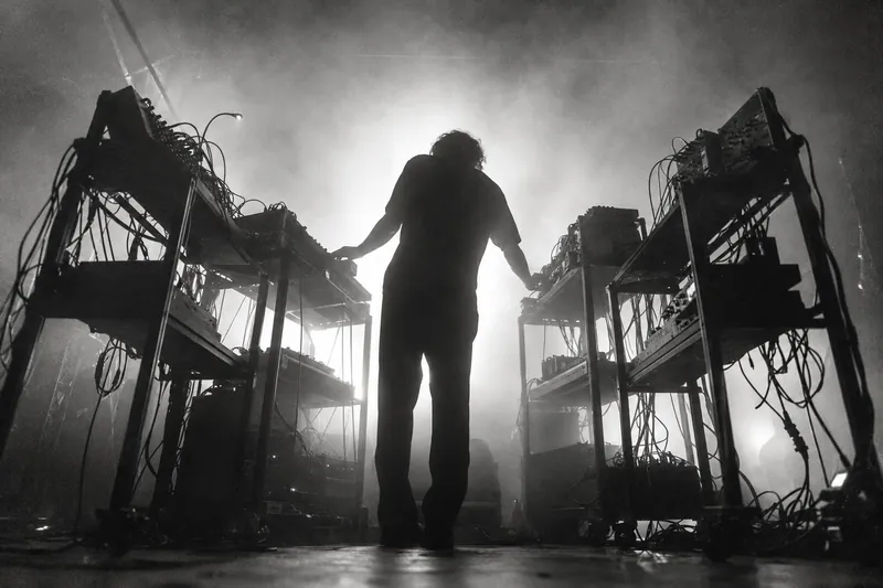 A musician silhouetted between two equipment stands of synthesizers and cables, in front of bright white backlight and haze.